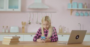 School preteen girl using mobile phone application for watching popular video online. Kid stays at home. Girl in pink shirt with cell phone sitting at pink kitchen.
