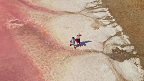 drone flying around lovely young couple walking together hand to hand along bright beautiful pink lake with salty shore. newlyweds tourists walking on white salty lake coast.