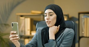 Young cheerful muslim woman in black traditional hijab talking by video via webcam on smartphone with client in office. Arabic female manager speaking with customer in videochat on phone Videochatting