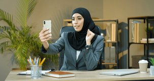 Beautiful muslim young woman in headscarf sitting at table in office and talking via webcam on smartphone. Pretty Arabian businesswoman in hijab having videochat on mobile phone. Videochatting.
