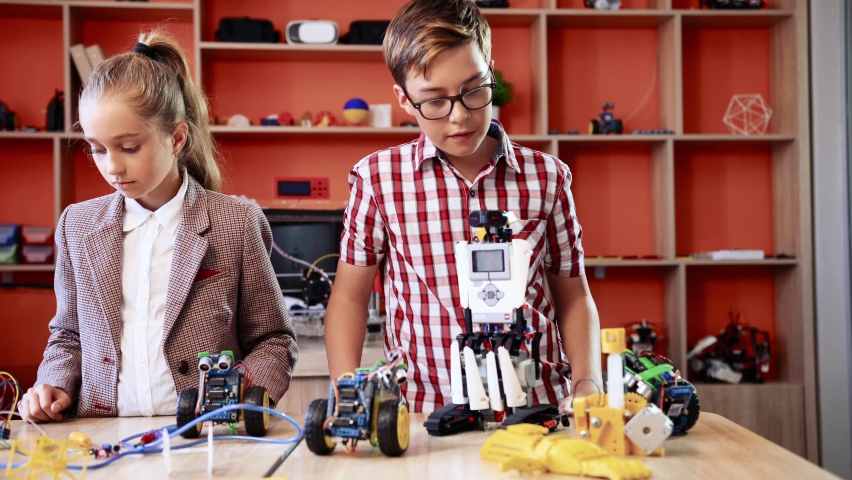 Portrait of two happy children standing at the table with robots. Beautiful girl and cute boy looking at each other smiling at camera while standing in robotics class.