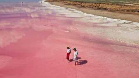 cirle drone flight over lovely couple tourists with corgi dog walking on bright beautiful landscape with beautiful pink lake and clouds reflections on water surface