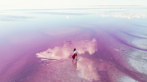 happy carefree young girl in red skirt and straw hat joyfully walking in water of amazing pink lake with calm surface and clouds reflections