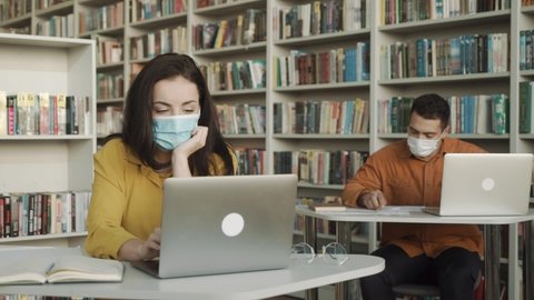 university students wearing face mask and sitting with books and laptop in library, social distance to avoid the spread of coronavirus in University.