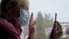 Ill old elderly woman at window in protecting medical mask on self-isolation. Communicate video link on smartphone phone with relative, waving hand. Family connection at quarantine covid-19 pandemic