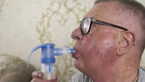 Portrait of a senior sick man breathing with the help of nebulizer. There is an ill old man with chronic obstructive pulmonary disease. He is sitting in a room and using an inhaler. Healthcare concept