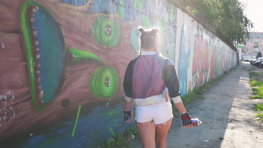 Concept of freedom and lifestyle. Happy girl walking and spraying aerosol paint into air. Creative teenager graffiti artist having fun at hot summer day against painted wall. Wild and free.
