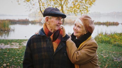 Happy retired couple, grandparents in warm elegant outerwear are talking, smiling and looking at each other with love while posing in city park against a river. Autumn day. Family relations. Close up