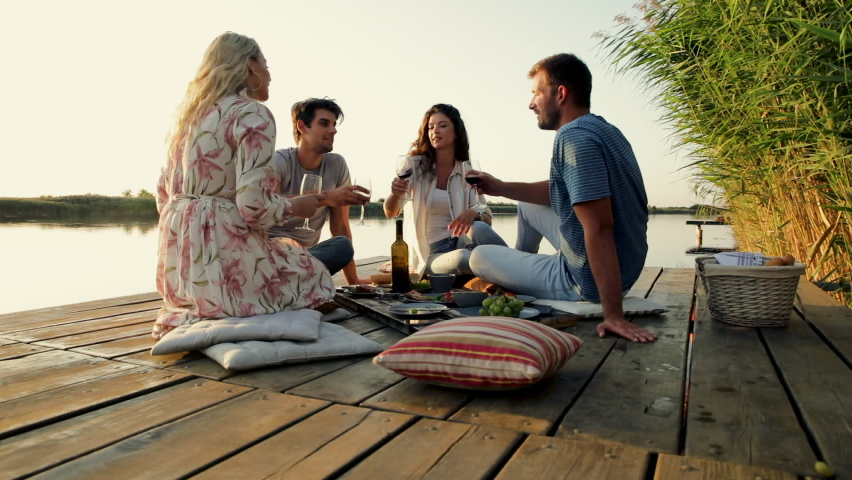 Group of friends having fun on picnic near a lake, sitting on pier eating and drinking wine. Royalty-Free Stock Footage #1062261319