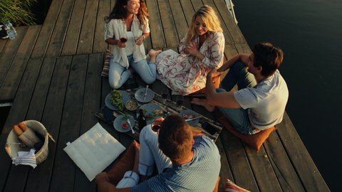 Group of friends having fun on picnic near a lake, sitting on pier eating and drinking wine. Stock Video