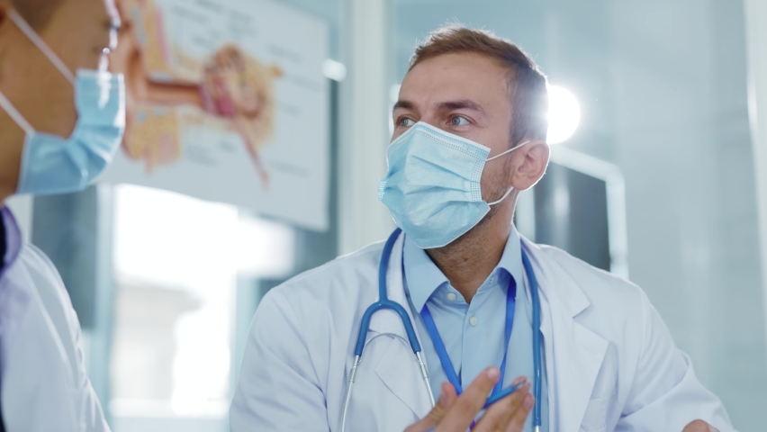 Multi-cultural men doctor and assistant wearing antiviral face masks talking consulting at hospital office discussing disease symptoms influenza. Medical couple. | Shutterstock HD Video #1062262096