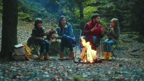 Family sits near campfire on autumn evening tells scary stories. Children with their parents are resting in the woods. Childhood relationship camping. Close up. Slow motion