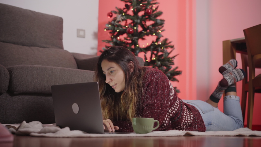 Smiling woman lying on the living room floor and making a video call with the laptop, celebrating Christmas alone and online staying at home. | Shutterstock HD Video #1062263302