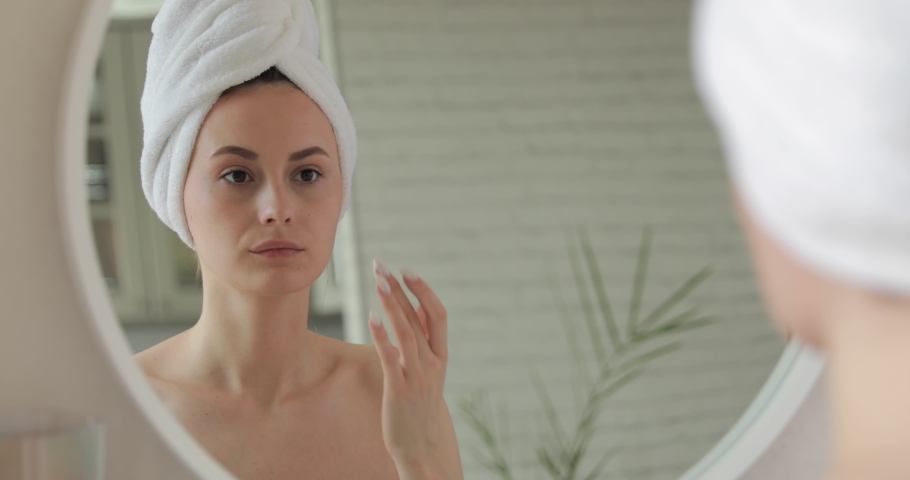 Happy lady in white towel standing in bathroom and applying cream on face. Young woman with bare shoulders using korean cosmetics for skin care. | Shutterstock HD Video #1062265039