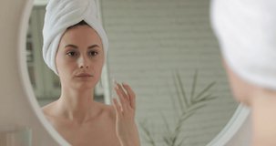 Happy lady in white towel standing in bathroom and applying cream on face. Young woman with bare shoulders using korean cosmetics for skin care.
