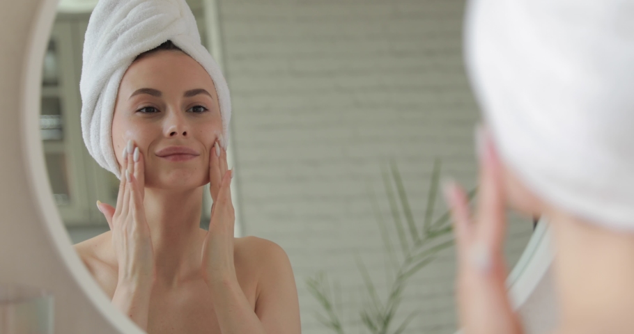 Happy lady in white towel standing in bathroom and applying cream on face. Young woman with bare shoulders using korean cosmetics for skin care. | Shutterstock HD Video #1062265039