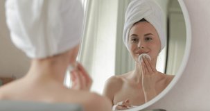 Beautiful female wrapped in white towel moisturising her skin during morning routine. Young woman with natural beauty using cotton pad at modern bathroom.