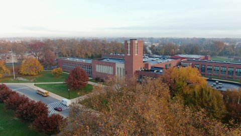 Aerial reveal of brick school building with school bus at front entrance. Drone flight over fall foliage, colorful leaves in autumn. American flag on pole in USA.