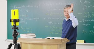 Asian senior male calculus professor is pointing on board while having blackboard lecture online through smartphone in the classroom at college