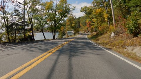 POV Driving a car on curvy asphalt road by the lake with colorful trees in fall season. Sunny day with sun beams