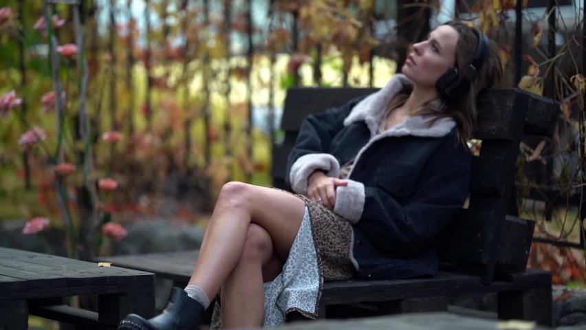 thoughtful young woman is sitting on bench at autumn day and listening to music by headphones, relax