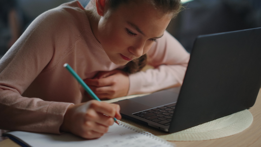 Portrait of focused child doing homework next to laptop computer at table. Teenage girl writing in notebook in front of laptop indoors. Closeup pretty schoolgirl studying at home education. | Shutterstock HD Video #1062279961