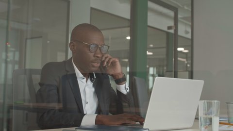 Medium shot of handsome African businessman wearing elegant clothes and eyeglasses is sitting at laptop in the office and talking on cellphone