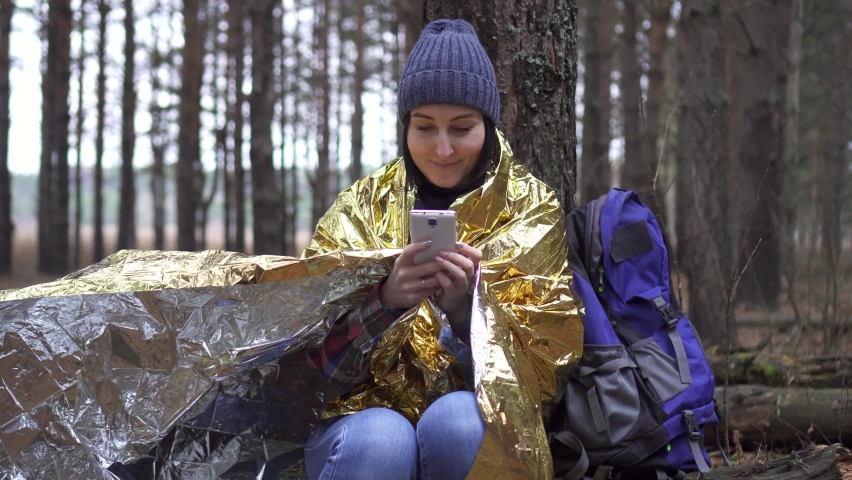positive woman tourist wrapped in a golden forest survival blanket uses a smartphone
