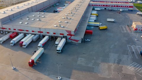 Aerial hyper lapse (motion time lapse) of the logistics park with a warehouse - loading hub. Semi-trucks with freight trailers standing at the ramps for loading/unloading goods at sunset. 