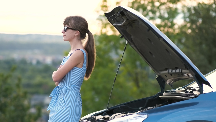 Young woman driver standing near a broken car with popped up hood waiting for help to arrive.