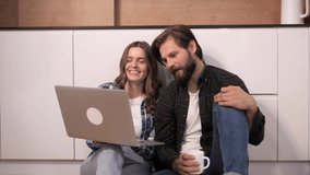 A happy couple in plaid shirts sitting on the floor in the kitchen watching movie make online purchases smiling hugging using laptop
