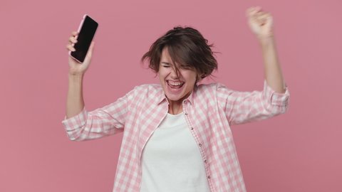 Smiling excited young brunette woman in casual checkered shirt posing isolated on pink background studio. People lifestyle concept. Talking on mobile phone say wow spreading hands doing winner gesture