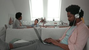 Afro man sitting on sofa with earphones on and using laptop while his family is sitting at the kitchen table near him
