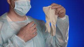 Doctor puts white latex protection gloves on his hands. Adult physician in surgical face mask, medical clothing on blue background. Concept virus infection control Omicron variant of SARS-CoV-2, COVID