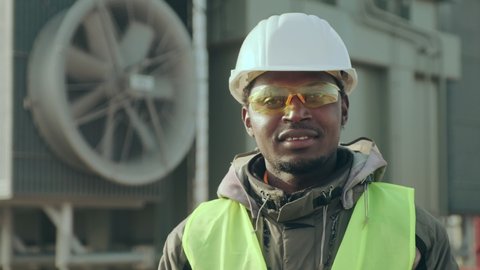 African-American construction engineer in reflective vest helmet and goggles stands at electric transformer substation fan closeup