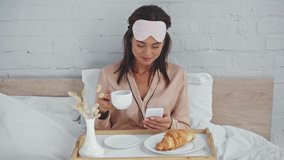 brunette woman drinking coffee and messaging on smartphone in bed