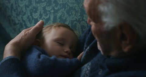 Cinematic close up shot of senior gray hair grandfather cuddling grandson baby is sleeping peacefully on arms while sitting on sofa at home. Concept:life, grandparents,love,care, generation, childhood