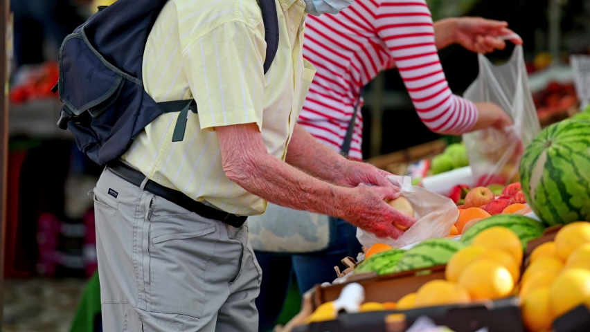 Elderly man wearing a protective face mask and shopping for oranges at an outdoor fruit and veg market stall.