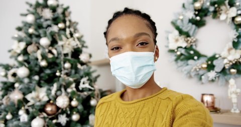 POV of a happy African American woman in masks isolated at home with a decorated Christmas tree with lights video chatting online and greeting friends and relatives with xmas. Xmas eve in quarantine.