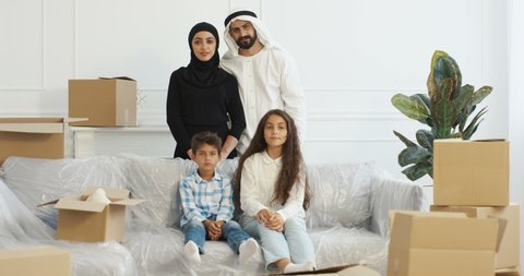 Portrait of happy muslim family smiling to camera and posing in living room. Moving in new accommodation. Arabian mother, father, daughter and son among carton boxes at home.