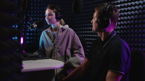 Professional radio recording studio. Beautiful DJ girl and young guy producer are recording a podcast. They create a radio broadcast. Dialogue together, discussing hot news