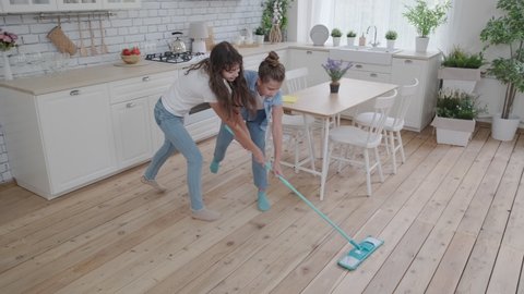 Funny girls hold a mop together and cheerfully clean the floors at home