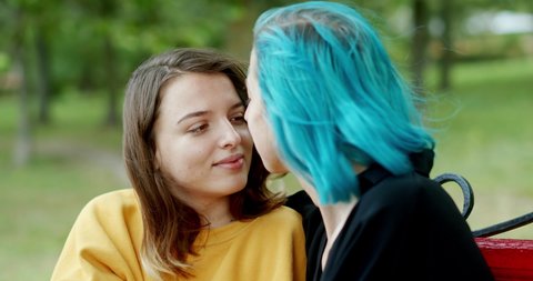 Young girlfriends embracing on a bench in tha park. Young woman with dyed hair hugging and kissing happy girlfriend while resting on bench on windy day in park.