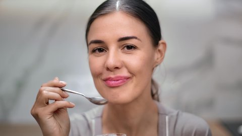 Closeup smiling face of domestic young woman eating yogurt use spoon at kitchen home having positive emotion. Portrait of happy female enjoying dairy breakfast or lunch. Shot with RED camera in 4K