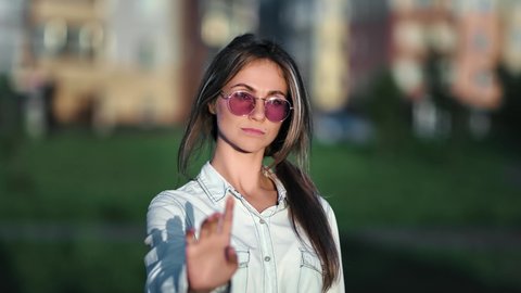 Portrait of confident trendy female in pink sunglasses shaking hand gesture no outdoor at summer city. Woman warning with admonishing finger disapproval sign. Medium close up shot on 4k RED camera