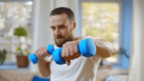 Portrait of young fit man looking at camera and exercising with dumbbells at home. Fitness trainer having online class working out at home. Handsome bearded guy training