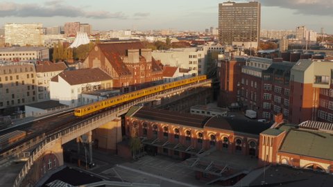 Typical Berlin Yellow Subway Train on high ground passing through city neighbourhood in beautiful golden hour Sunset light, Aerial tracking follow shot wide angle