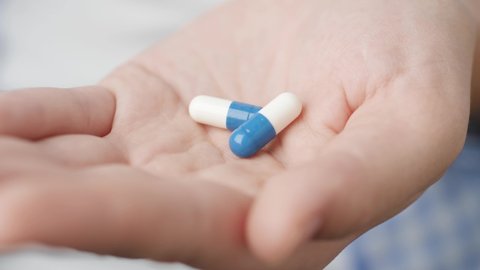Two big white-blue cylindrical capsule pills fall into palm of hand from pill bottle. Close-up, front view, center composition