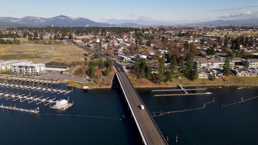 Traffic bridge crossing the beautiful Spokane River into Post Falls Idaho with trees blue sky businesses and homes with mountains in the background.