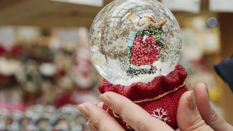 Close-up of snowy Christmas globe toy in female hands. Woman shopping gifts in Christmas fair